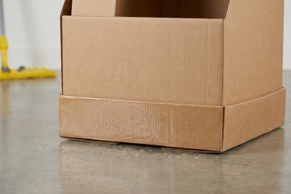 Base de boîte en carton humide Les dix centimètres inférieurs d'une boîte en carton ondulé présentent une absorption d'humidité importante provenant d'un sol en béton mouillé, ce qui met en évidence la capacité du papier non traité à absorber l'eau.