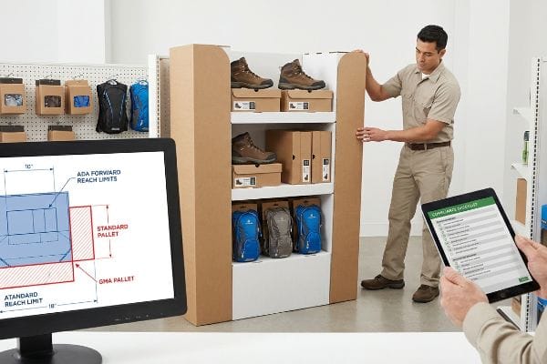 Brown corrugated cardboard display assembly with hiking boots and backpacks, a monitor showing ADA Forward Reach Limits, and a tablet with a Compliance Checklist.