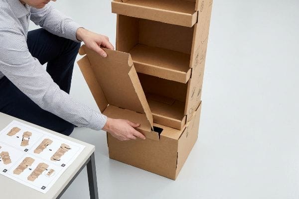 A person assembles a brown cardboard display stand, inserting a flap into a slot, with assembly instructions visible nearby.