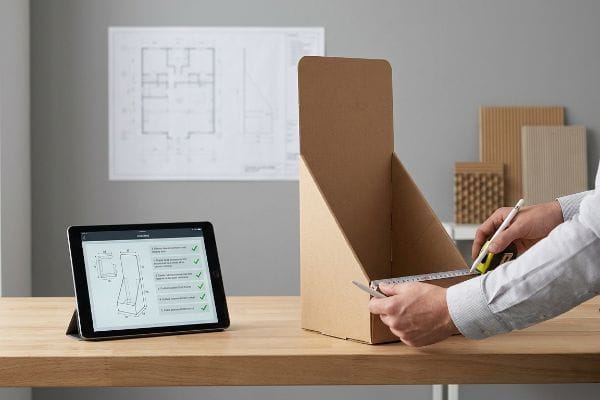 Brown corrugated cardboard display being measured with a utility knife and tape, alongside a tablet showing a display design and a blueprint.