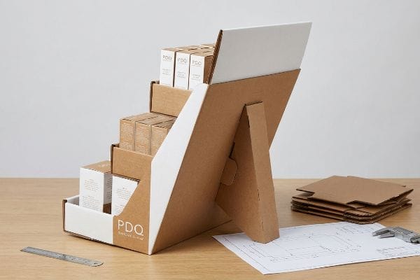 Présentoir de comptoir PDQ en carton kraft avec support chevalet, contenant des produits en boîte blanche, à côté d'un gabarit et d'un pied à coulisse.