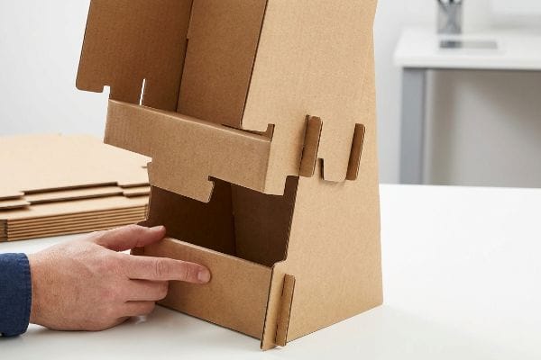 Brown corrugated cardboard display stand assembly, a hand interlocking a shelf into the main structure with visible slots and tabs, highlighting testliner components.