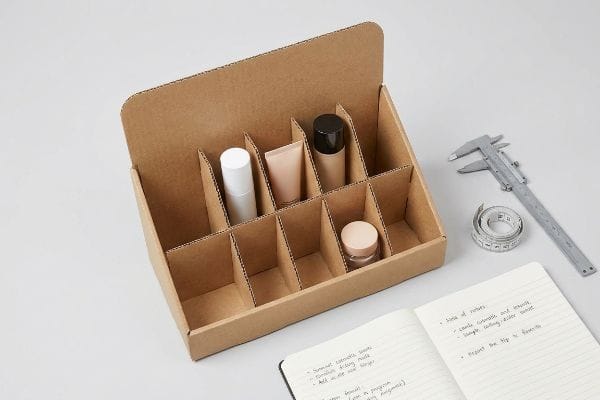 Cardboard Display Prototype Sample A brown corrugated cardboard counter display prototype holds various cosmetic products, alongside a caliper, measuring tape, and a notebook, highlighting dimensional validation.