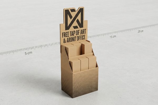Corrugated cardboard point-of-purchase display featuring a black 'M' logo and 'Free Tap Of Art & Aronit Office' text, holding six small brown boxes on two tiers.