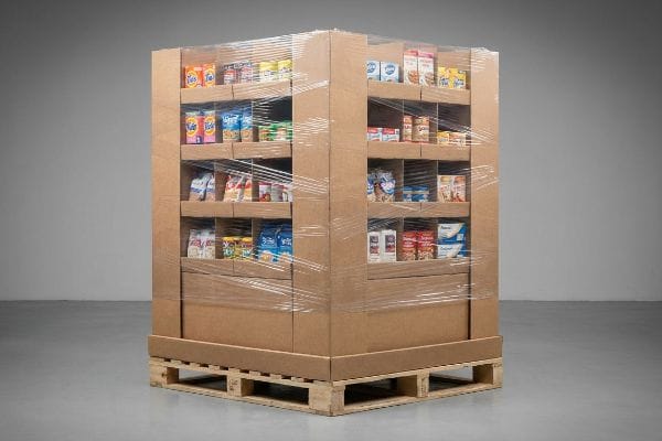 A fully kitted corrugated cardboard endcap display, loaded with diverse grocery products and secured with clear shrink wrap, rests on a wooden shipping pallet.