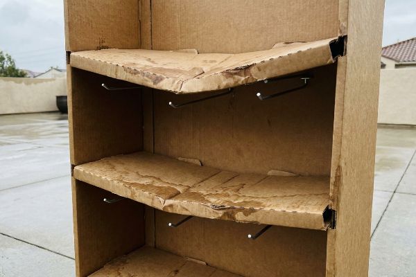 Water-damaged cardboard display with warped shelves and bent metal hooks, showing structural failure from outdoor exposure.