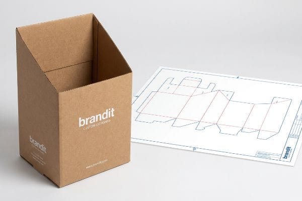 An empty Brandit custom designed cardboard dump bin stands beside its die-line blueprint, showcasing structural engineering for retail displays.