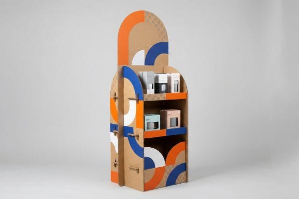 Cardboard pop-up display stand features bold orange, blue, and white geometric patterns, holding small product boxes.