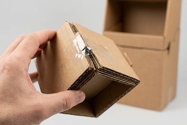 Corrugated cardboard box with a hand holding a taped folding tab, revealing a micro-tab failure and visible flute thickness.