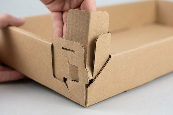 茶色の段ボール製のPDQディスプレイを組み立てている人の手。小売店向けの包装のために、角のロックタブを固定している。.