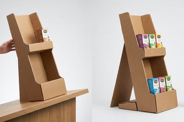 Mostrador expositor de dos niveles de cartón corrugado marrón con soporte trasero tipo caballete, que se muestra vacío y con cajas pequeñas de productos.