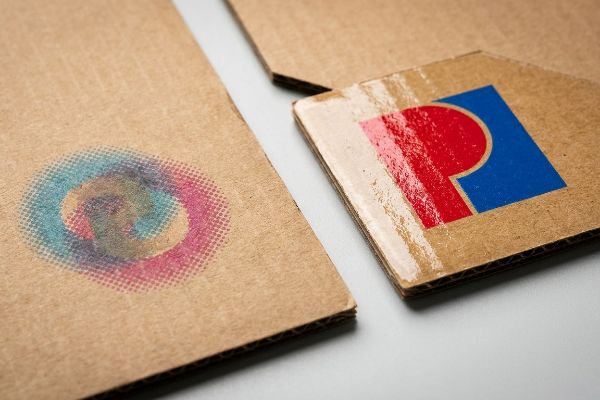 Raw corrugated cardboard displays faded CMYK halftone dots next to a vibrant red and blue spot color logo, highlighting testliner printing.