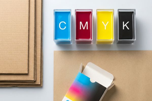 Arrangement of CMYK (Cyan, Magenta, Yellow, Key) ink containers, stacked corrugated cardboard, and an open retail packaging box.