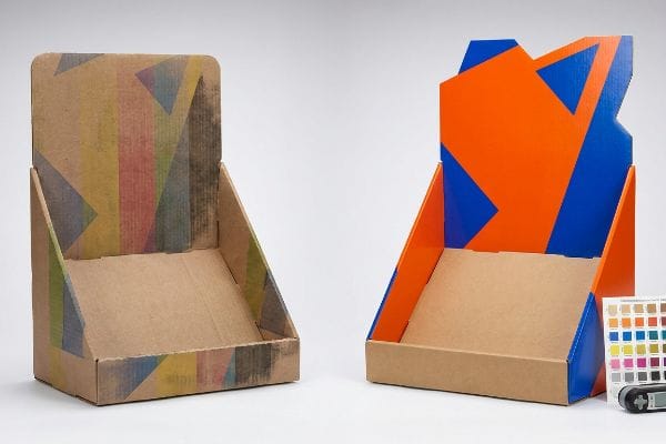 Two corrugated cardboard display stands, one with muted CMYK geometric print, the other with vibrant orange and blue Pantone spot colors and a die-cut header.