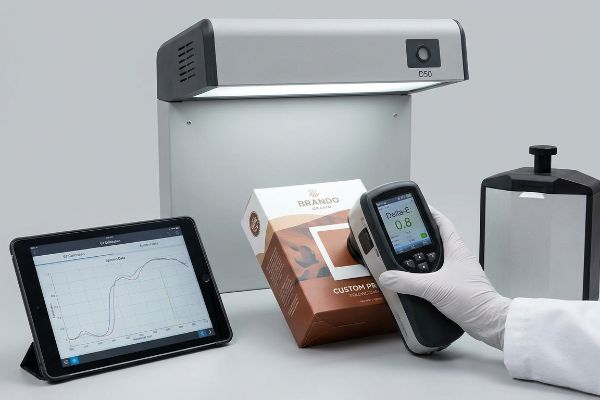 D50 light booth, Brando Original Custom Print box, spectrophotometer showing Delta-E 0.8, and a tablet with spectral data.