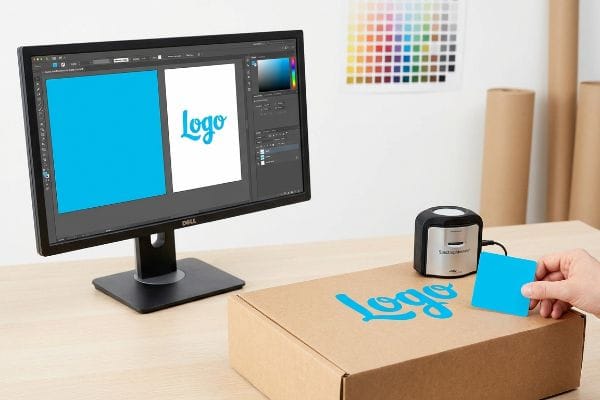 A designer uses a ColorChecker device to match the blue Logo on a cardboard box with the digital artwork displayed on a Dell monitor, ensuring accurate print color.