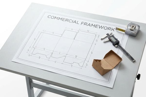 A blueprint for a cardboard box dieline, labeled 'Commercial Framework,' lies on a drafting table with a digital caliper, tape measure, and a small brown cardboard box.