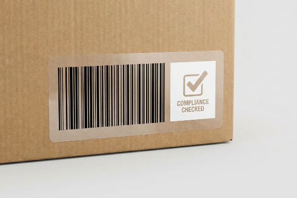 A brown cardboard box displays a white label with a black barcode and a 'Compliance Checked' stamp.