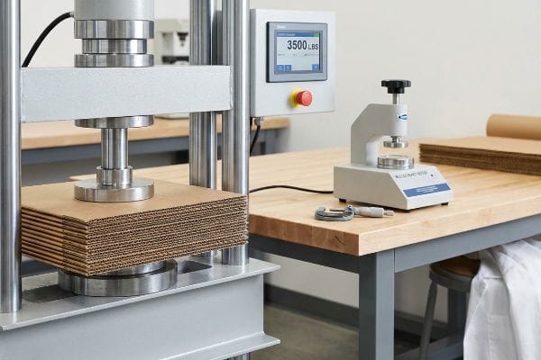 Corrugated cardboard stack under a hydraulic press showing 3500 Lbs, beside a Mullen Burst Tester and micrometer.