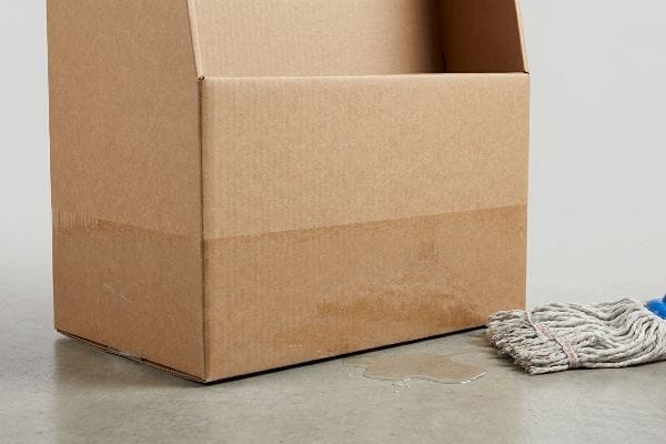Brown corrugated cardboard box base absorbing water from a floor puddle, illustrating moisture wicking vulnerability, beside a wet mop.