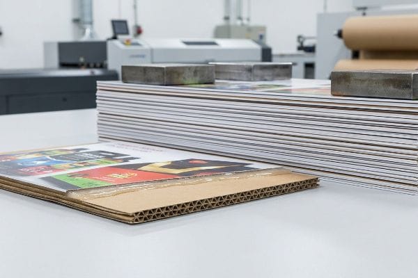 Corrugated display boards with colorful printed top-sheets stacked in a factory, showing the exposed fluting and metal weights for curing.