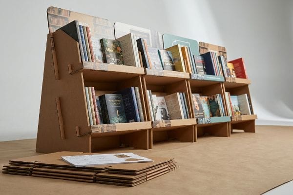 Corrugated cardboard book display stand filled with books like 'The Main' and 'Forgotten Towers', with flat-packed components.