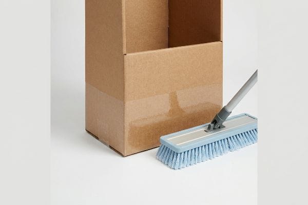 An open brown corrugated cardboard box stands next to a light blue industrial mop with a grey handle.