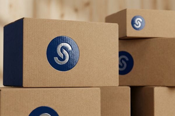 Une pile de boîtes en carton ondulé arbore un logo circulaire bleu bien visible avec un « S » argenté sur chacune, illustrant la qualité supérieure des matériaux utilisés en PLV.