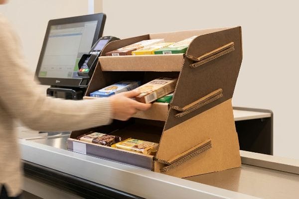 A shopper's hand reaches for a product on a multi-tiered corrugated cardboard POS display at a retail checkout.