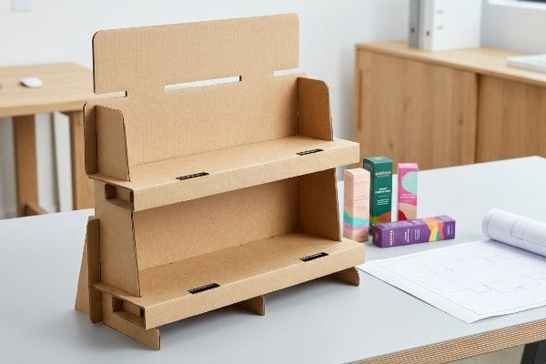 Two-tier corrugated cardboard point-of-purchase display on a desk with colorful product boxes and a blueprint.
