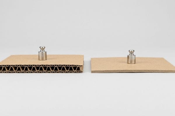 Corrugated cardboard with visible flutes and solid chipboard, each bearing a metal weight, illustrating structural load capacity.