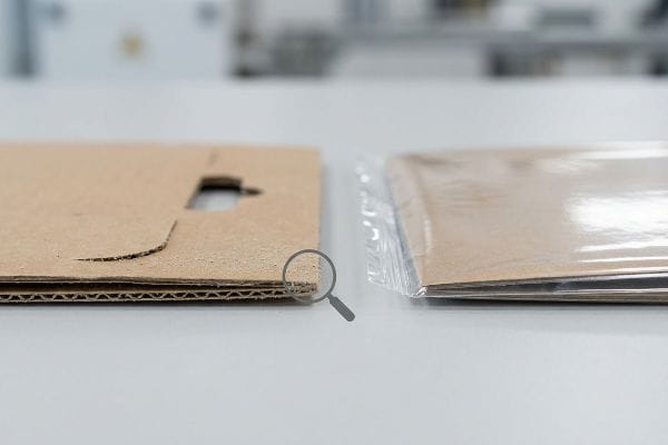 Close-up of raw corrugated cardboard showing visible paper dust and fractured fibers on die-cut edges, next to a clean, plastic-wrapped laminated board.