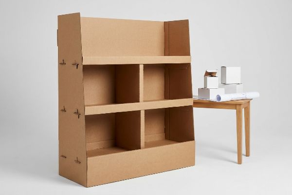 An assembled prototype floor display made from unprinted corrugated board, with white boxes and a blueprint on a table.