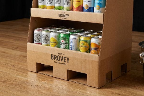 Brown corrugated display, The Brovey Paper Boret, holds cold beverage cans with condensation on a wet wooden floor.