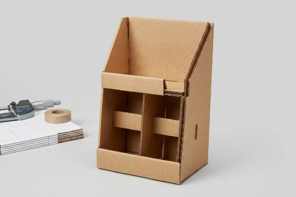 Brown corrugated cardboard display with internal H-dividers, next to a digital micrometer, tape, and dieline blueprints.