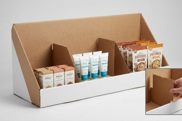 Brown corrugated kraft paper countertop display with white front and modular dividers, organizing Spireale boxes, Fioré creams, and Protenoes pouches.