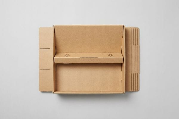 An unassembled cardboard book display stand features a partially folded brown corrugated board with a shelf insert showing down arrows, alongside stacked flat components.