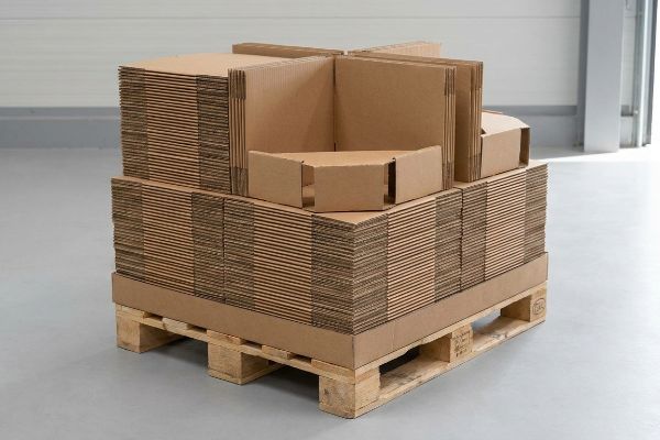 A large stack of flat-packed corrugated cardboard sheets and partially assembled display components rests on a wooden pallet.