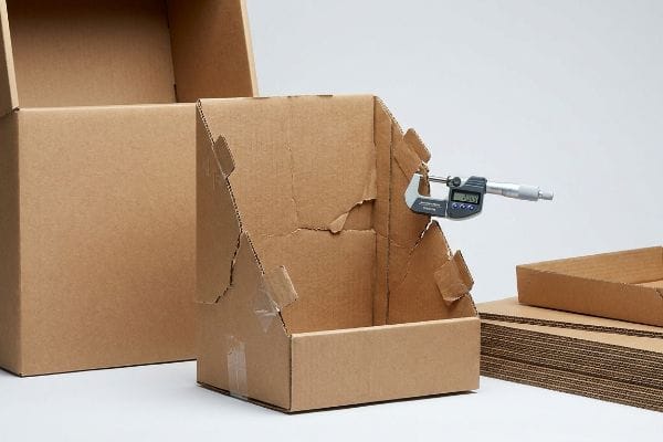 A digital micrometer measures B-flute corrugated cardboard on a poorly folded display unit, revealing cracking and clear tape.