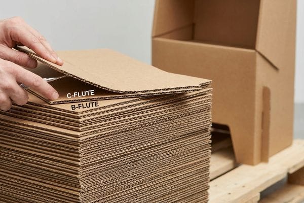 Hands separate corrugated C-Flute and B-Flute cardboard sheets, revealing their distinct fluting from a large stack, with an empty box nearby.