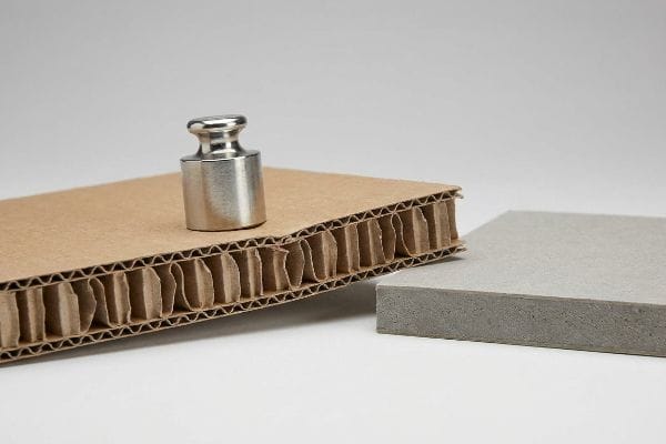 Corrugated cardboard with visible fluted arches supporting a metal weight, next to a solid grey board.