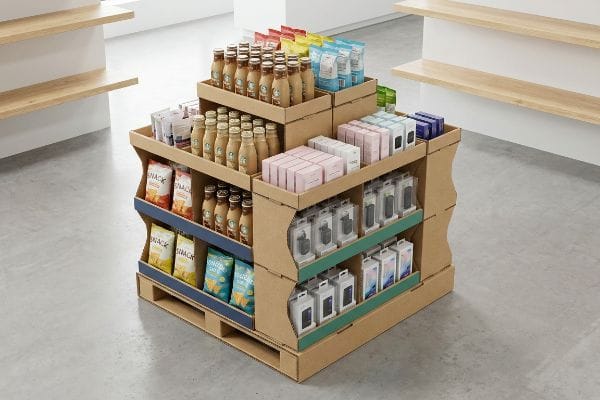 Présentoir en demi-palette ondulée dans une allée de magasin, présentant des boissons en bouteille, des sachets de collations emballés et des appareils électroniques en boîte avec des accents bleus et verts.
