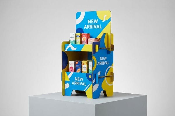 Un présentoir PLV en carton ondulé bleu et jaune vif met en valeur les nouveaux produits sur deux étagères, avec des formes abstraites et l'inscription « Nouveauté ».