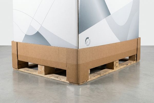 Brown corrugated pallet wrap encircling a large white retail display box on a wooden pallet, protecting its base.