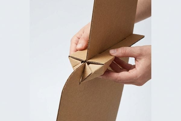 Natural brown corrugated cardboard pieces are interlocked by hands, forming a precision paper lock joint for a display stand.