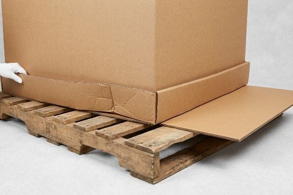 غطاء من الورق المقوى المموج يسد الفجوات الواسعة على منصة خشبية، لحماية صندوق كرتوني كبير من الترهل، مع يد ترتدي قفازًا تقوم بتعديله.