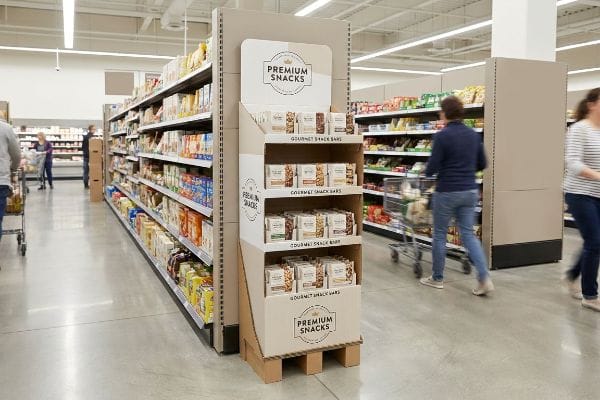 Kunden schlendern durch einen Supermarktgang, in dem ein Bodenaufsteller aus Wellpappe für „Premium-Snacks“ und „Gourmet-Snackriegel“ wirbt.
