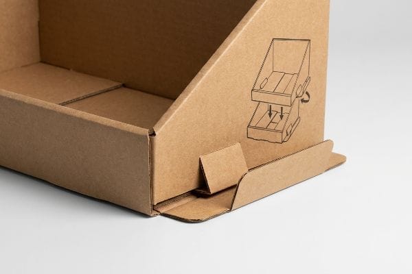 Brown corrugated cardboard PDQ display tray featuring a printed assembly diagram for its auto-locking crash bottom.