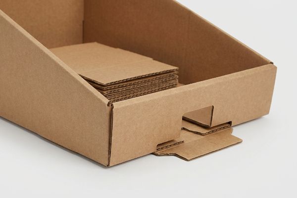 Brown corrugated cardboard PDQ display tray with a visible pre-glued crash-bottom structure, containing stacked flat cardboard inserts.
