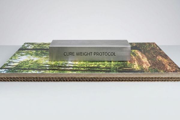 Brushed metallic block engraved 'Cure Weight Protocol' applies dead-weight pressure to litho-laminated corrugated board with a forest print.